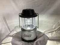 Front view of silver and black Coleman battery operated lantern with clear plastic globe, handle, and label.
