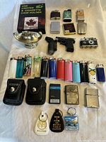 Photo of lighters and key tags arranged on white fabric. Items include disposable lighters, novelty shaped lighters, USB lighter in packaging, Zippo lighters with leather cases, mini torch shaped like handgun, and key tags.