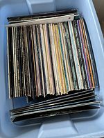 Photo shows a blue plastic tub filled with approximately 80 vinyl records with visible record spines in various colors and designs.