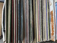 Photo showing side view of approximately 85 vinyl record covers stacked tightly in a box, showing varying colors and signs of wear on edges.