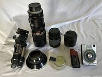Photography equipment consisting of four camera lenses of various sizes and brands, a colored lens filter, lens cleaner, metal lens hood, a red and black lens cleaner, compact adjustable mini tripod and small lens cap or case on a white fabric background.