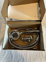 Photo showing chrome finish shower head with multiple spray nozzles, a matching flexible coiled metal hose, and a metal plumbing assembly piece inside an open cardboard box.