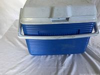 Blue Rubbermaid cooler with white lid and handle placed on a white surface.