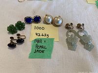 Five pairs of earrings arranged on white fabric with handwritten notes showing weight and materials