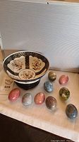 Photo of hand-painted decorative bowl surrounded by ten polished stone eggs in various colors and patterns.