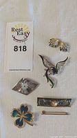 Photo of all jewelry laid out including clip-on earrings, bird brooch, flower brooches and bar pins on a beige fabric background with seller's tag visible.