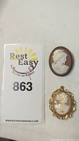 Two cameo brooches shown with auction lot number card, one brooch features a gold ornate frame, both have white carved female profiles on brown background.