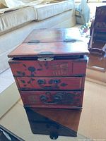 Closed view of antique wooden jewelry chest with faded red and black paint with metal drawer handles and painted characters on front.