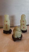 Photo of three jade figurines displayed on a surface with a background of horizontal lined texture. The two standing figurines hold objects with red detail, mounted on dark wooden stands. The smaller seated figurine is also on a wooden stand and positioned in front.