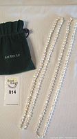 Two long strands of white pearls with a drawstring pouch labeled 'KAI YIN LO' placed on fabric background.