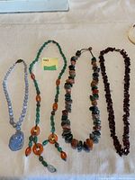 Four stone necklaces laid out flat on a cream fabric surface, showing varied bead shapes and colors from left to right.