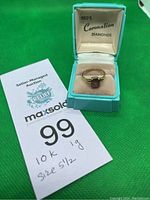 Gold ring with a purple rectangular stone displayed in a light blue ring box with a cream velvet cushion. The box lid shows the brand 'REG'S Coronation Diamonds'. A paper tag with auction info and handwritten note '10k 1g size 5 1/2' is visible.