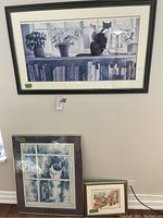 Wall hanging art of a cat sitting on a windowsill with potted plants and bookshelf below, framed with black molding and white mat