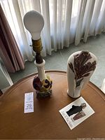 Photo of whole lamp and signed vase with artist card displayed on wooden table near window curtains.