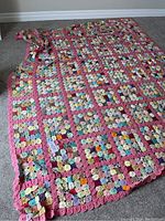 Full view of rectangular crochet bed cover featuring multiple colorful circular motifs joined by pink lattice borders, spread on carpet.
