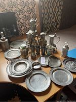 Overview of the entire pewter collection on a table including plates, pitchers, tankards, mugs, and the kettle with flower design visible.