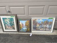 Three framed original paintings by Richard Wolff leaning against a garage door. Two smaller paintings on the left and a larger painting on the right.