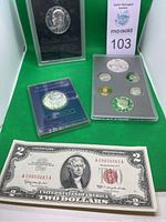 Four currency and commemorative items displayed against green background: red seal note, silver round, medallion and proof set.