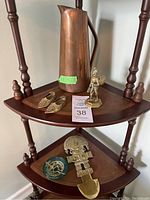 Photo shows large copper pitcher, brass figurine, pair of brass shoe figurines on a wooden shelf