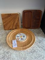 Three vintage Dansk pieces with two square teak boards at the back and one round wooden platter with decorative ceramic center in front.