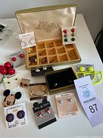 Overview photo showing vintage gold-tone jewelry case with multiple compartment sections, and several pairs of earrings in a variety of styles and colors arranged around it on a white surface.