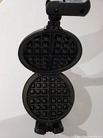 Open waffle maker showing non-stick grids with deep square pockets forming thick Belgian-style waffles in oval shape.