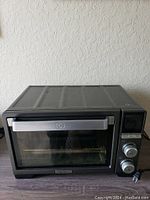 Front view photo of black Calphalon toaster oven with stainless steel handle and control knobs, placed on a wooden surface against a textured wall.