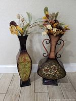 Two decorative floor vases side by side, both approximately 24 inches tall, with artificial flowers inside. The left vase is tall and slender with green and gold floral patterns. The right vase is wider with curved handles and painted floral motif. Both floral arrangements have autumn colors.