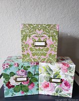 Front view of three floral file storage boxes arranged in a pyramid shape showing different floral prints and metal label holders.