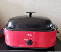 Front view of red Oster electric roaster oven with black dome lid and front temperature control knob.