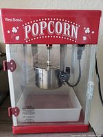 Red West Bend popcorn machine with clear acrylic panels, stainless steel kettle in the center, illuminated POPCORN text on top section, white tray at bottom.