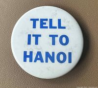 Front view of the pin showing white background with 'TELL IT TO HANOI' in blue block letters.