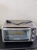 Black+Decker toaster oven closed with power cord on top, showing stainless steel finish and dials.