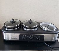 Photo of triple burner Crock Pot slow cooker with two ceramic pots with lids on base, one pot missing.