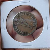 Front side of the token showing 'SUCCESS TO THE FISHERIES' and fish image, held in a white cardboard holder.