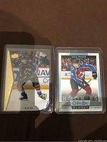 Two Cale Makar rookie cards propped upright against a dark surface. Left card is a die cut with gold foil accent and RC logo bottom right. Right card is a glossy O-Pee-Chee card with Makar skating, labeled 'ROOKIES'.