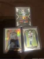 Three football rookie cards of Justin Herbert in protective sleeves: Select Club Level, Select Premier Level, and Mosaic Straight Fire Silver Prizm insert cards.