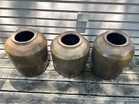 Three brown-green glazed ceramic planters arranged side by side on wooden deck against house siding