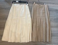 Two skirts side by side on hangers flat on floor showing off-white on left and beige on right, pleats and waistband visible