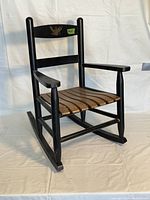 Side angled view of the antique children's rocking chair in black paint with natural wood slatted seat and eagle decoration on the backrest