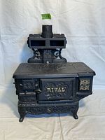 Front view of black cast iron RIVAL toy stove showing embossed floral details, chimney, and gold accents on front and side panels.