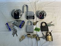 Photo showing nine padlocks including two circular disc locks, one open Master lock, two Loxon laminated steel locks with blue tags, one gray Sargent & Greenleaf lock, one black Abus lock, and three small brass locks connected by a chain, all with keys.