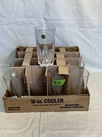 Front view of 12 lead crystal glasses arranged in original cardboard box, one glass lifted showing its size and shape, sticker label visible on glass.