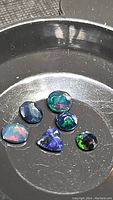 Six faceted Ethiopian black opal gemstones showing rainbow flash colors in round, oval, and triangular shapes in a black dish.