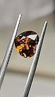 Close-up image of 1.97ct faceted pear-shaped orange zircon gemstone held in tweezers showing transparency, cut, and internal clarity.