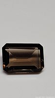 Single rectangular emerald cut natural smokey quartz gemstone with rich brown hue on plain background.