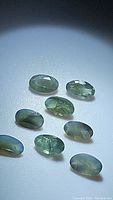 Eight oval greenish oval cut alexandrite gemstones with visible internal inclusions on white surface, illuminated by a focused light source.