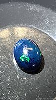 Close-up photo of an oval black opal cabochon gemstone showing vivid green and blue light flashes on a dark base color.