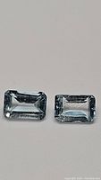 Two natural sky blue topaz gemstones, emerald cut, laid flat on a white surface. Stones are transparent with visible reflections and rectangular shape.