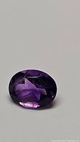Close-up image of an oval cut deep purple amethyst gemstone placed on a gray background, showing facets and clarity.